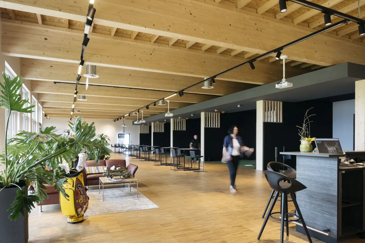 Das Indoor Golfstudio Götzis: Lounge mit Ledersofa und Golfbag, Stehtische, die Bar und rechts die Simulator-Boxen unter der Holzdecke
